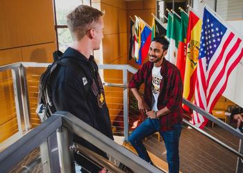 Two students in the student center talking on the steps to second floor with a bunch of international flags behind them.